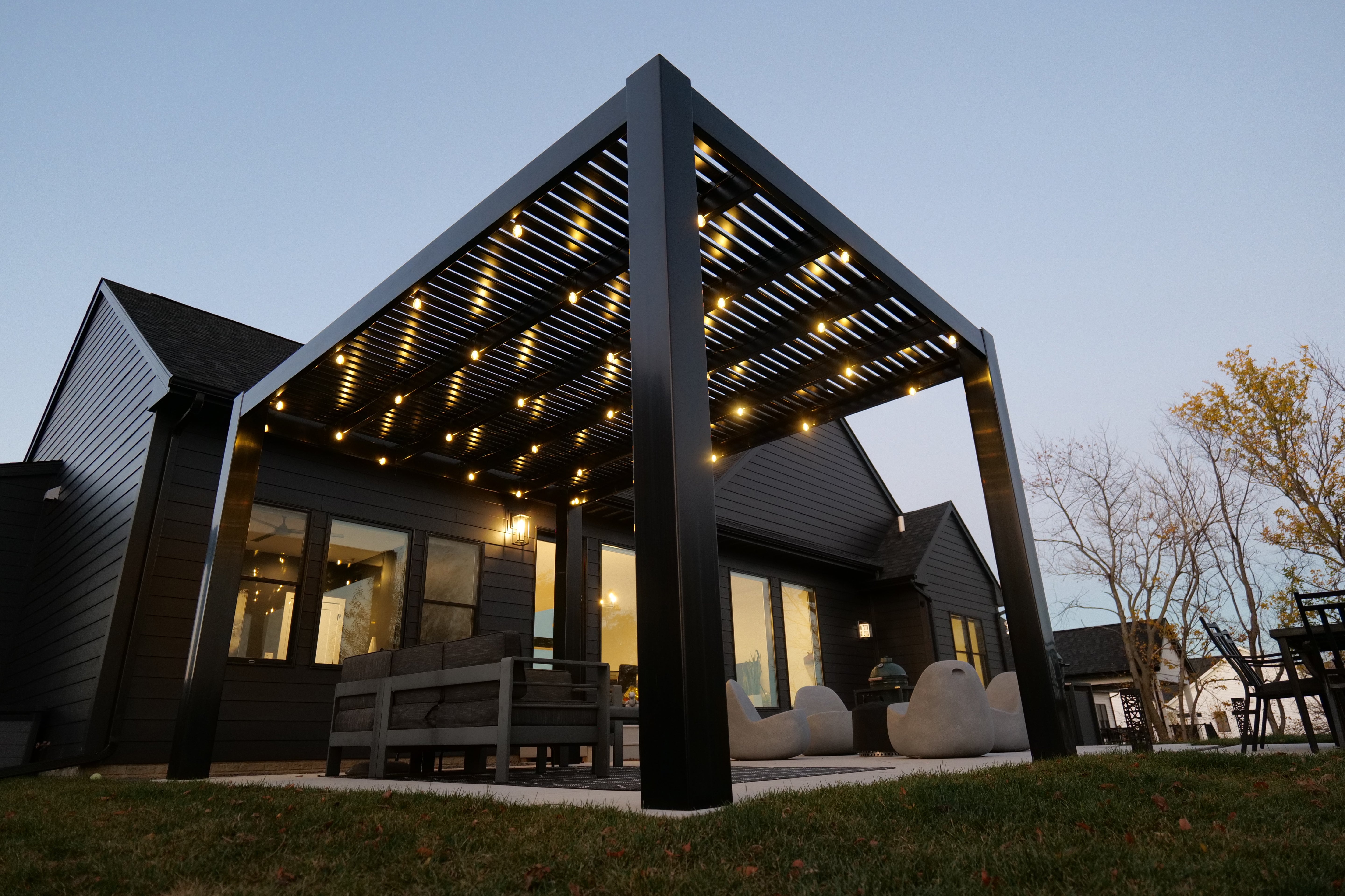 Modern outdoor pergola by Moda Pergolas with string lights on a house exterior at dusk.