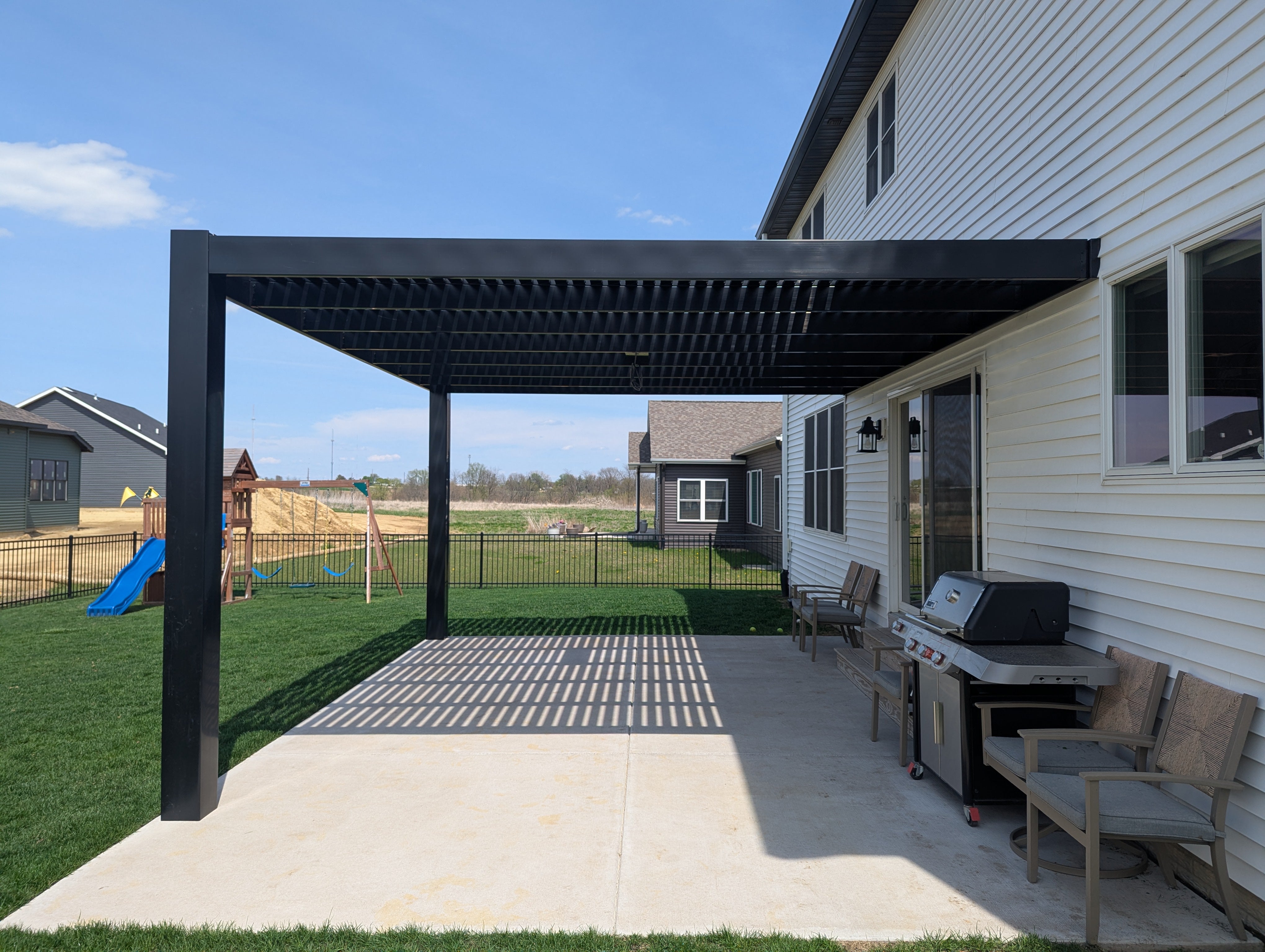 Outdoor patio with black attached modern pergola, grill, and chairs on a sunny day.