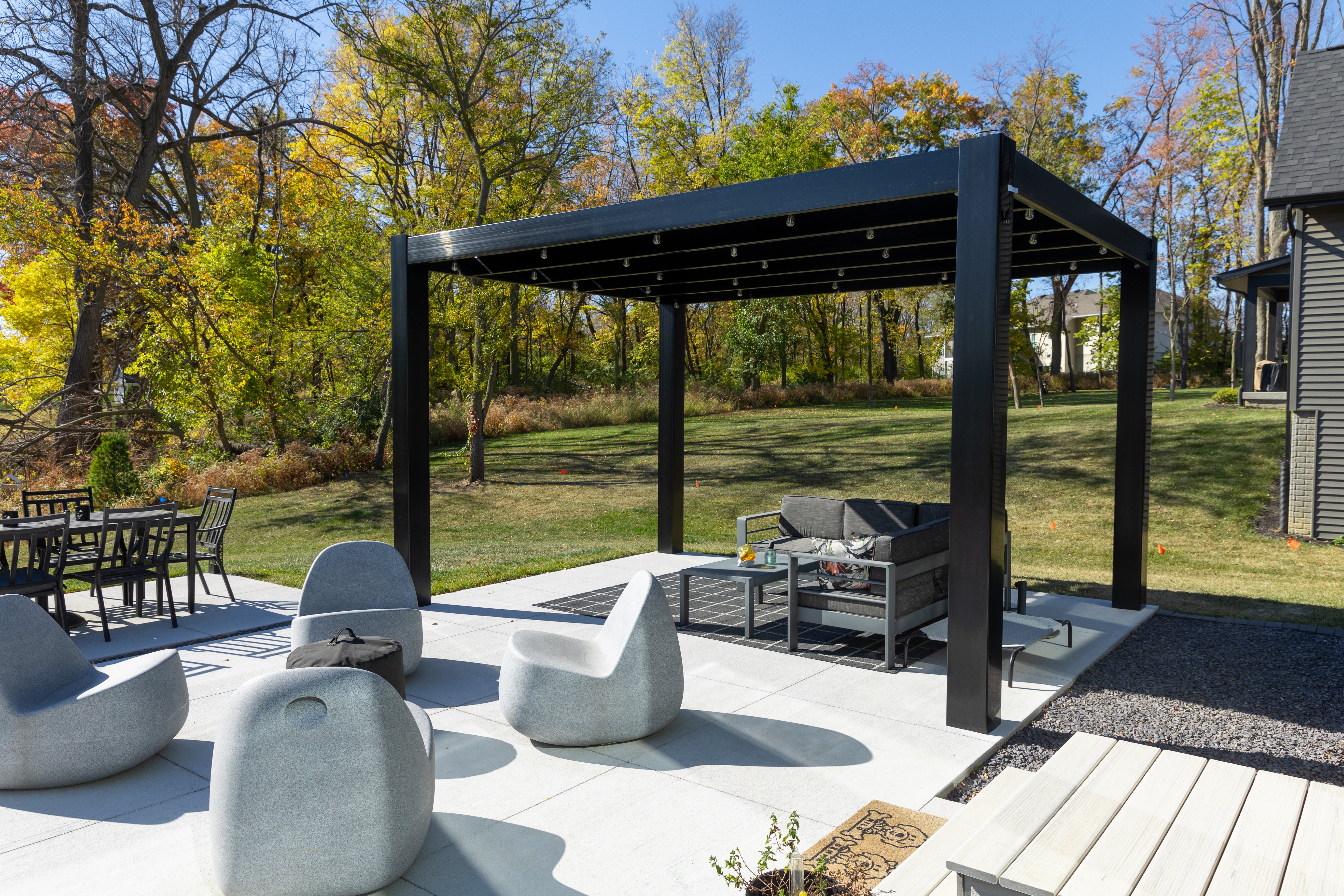 Outdoor patio with black modern pergola by Moda Pergolas, outdoor furniture, and trees in the background
